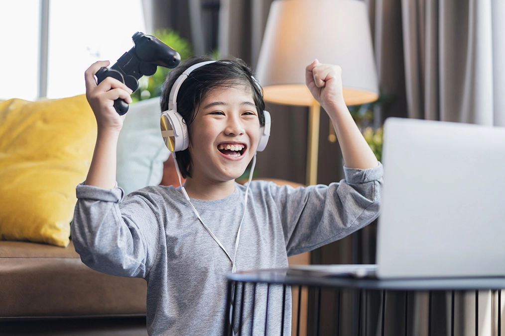 Asian child boy wear headphone hand playing laptop game online with exited and fun sitting on floor at living room at home, son is playing alone with mobile equipment.Amazed happy schoolboy raising arms and smiling while sitting on sofa celebrating victo