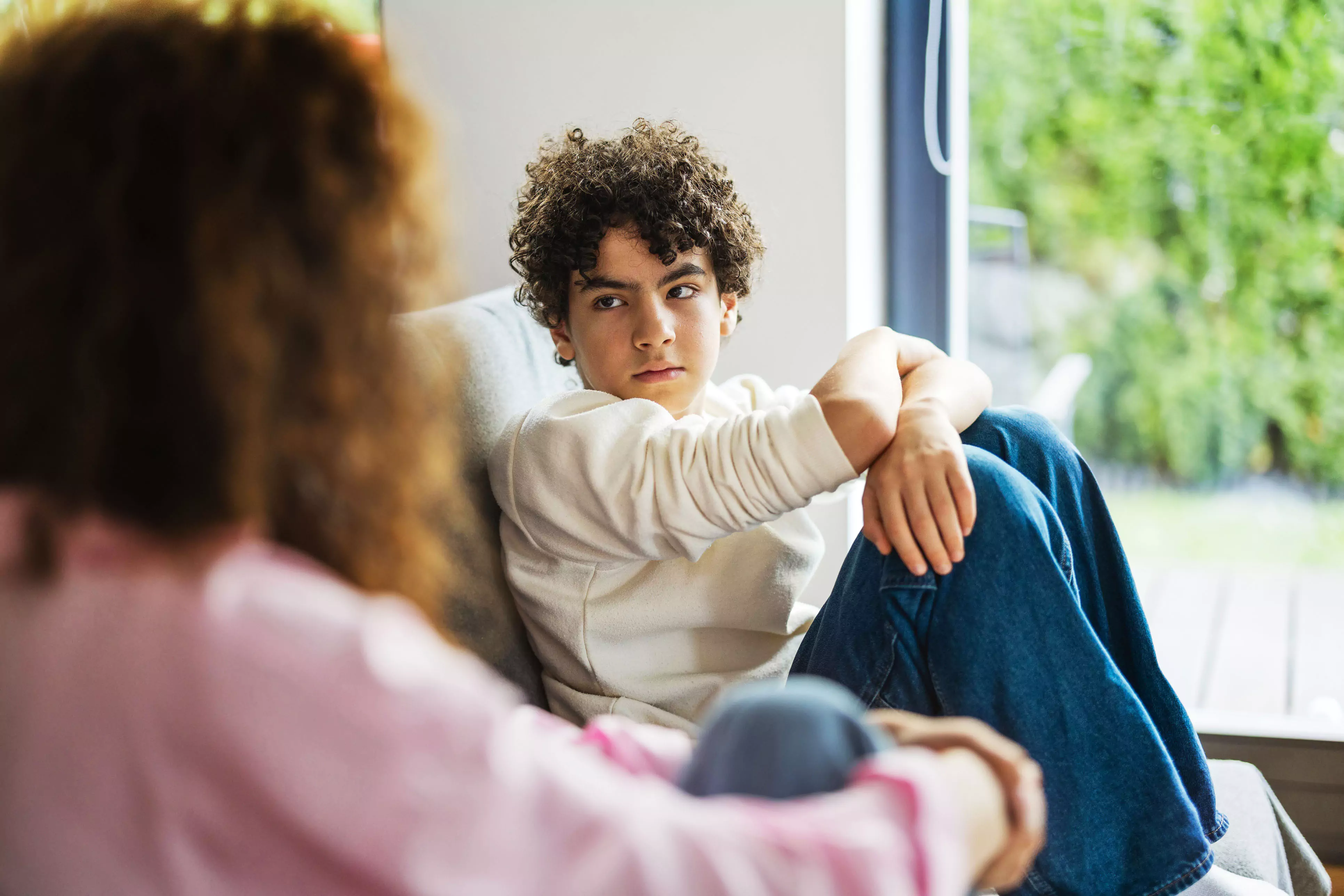 Teen boy sits on couch, knees drawn up to chest, with worried look on his face.