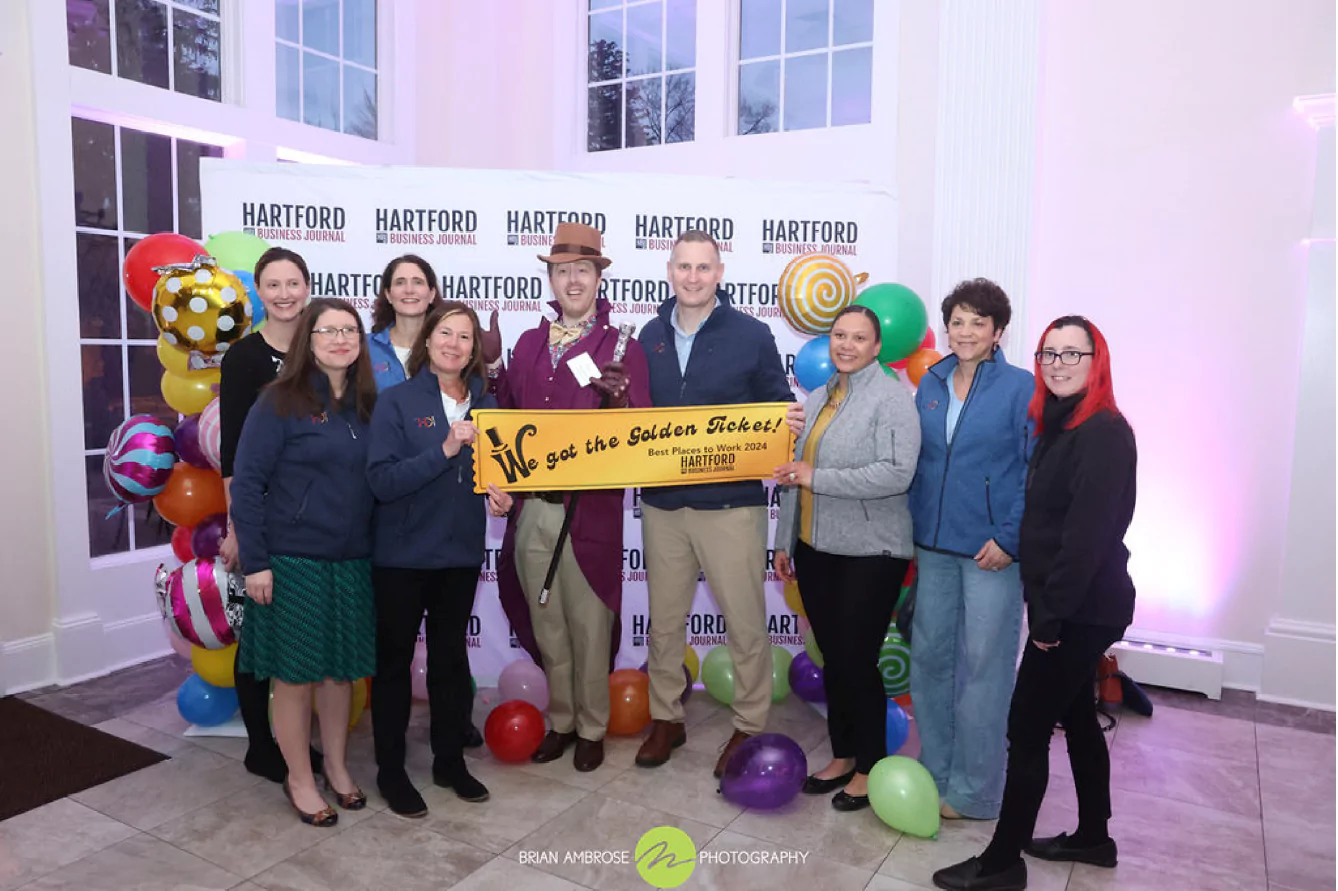 A group of CHDI staff pose together at the 2024 Best Places to Work awards dinner. The event was Wonka-themed, so they are holding an oversized golden ticket and standing with the emcee who is dressed as Willy Wonka.