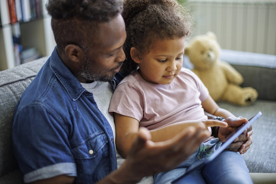 Dad and young daughter sit together on couch looking at iPad together