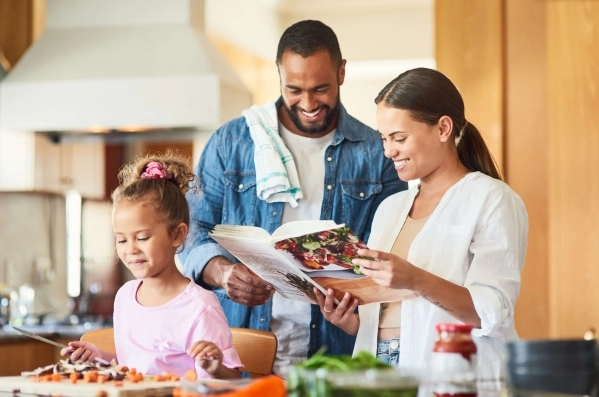 Two parents and a young girl look at a cook book together and chop food in a kitchen.