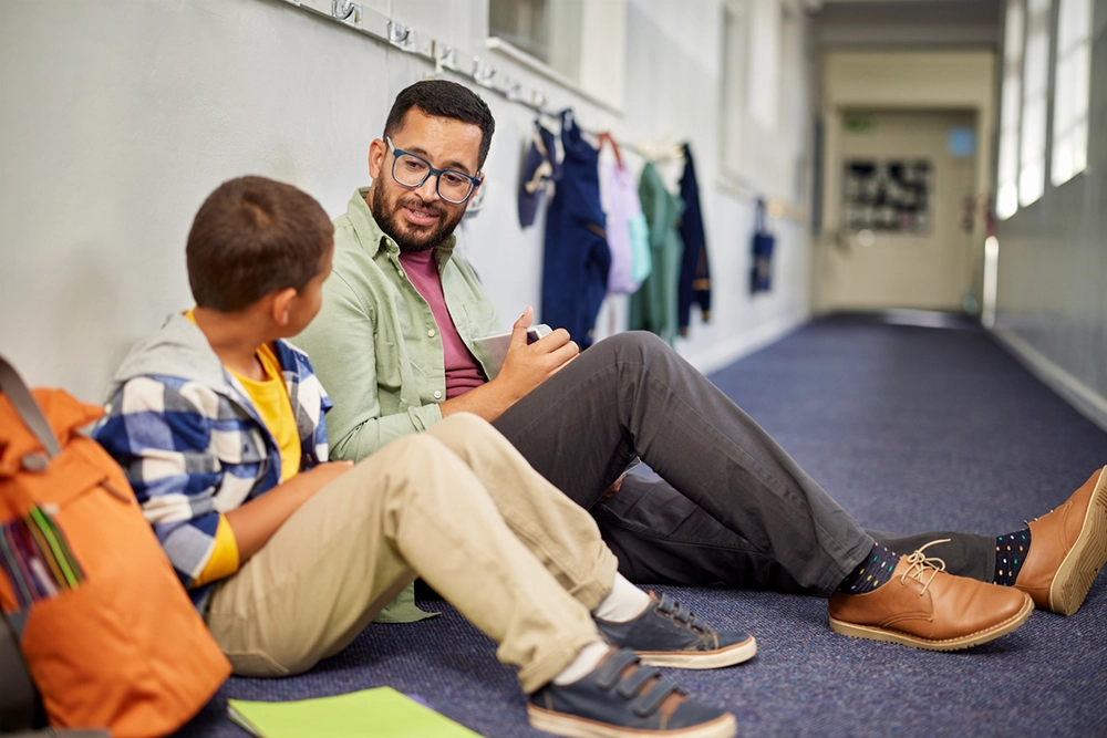 Young male psychologist speaking to depressed boy in school while sitting on the floor in school corridor. Man working with a child while discussing about learning problem and bullying. Young and friendly teacher speaking to boy in school hallway.