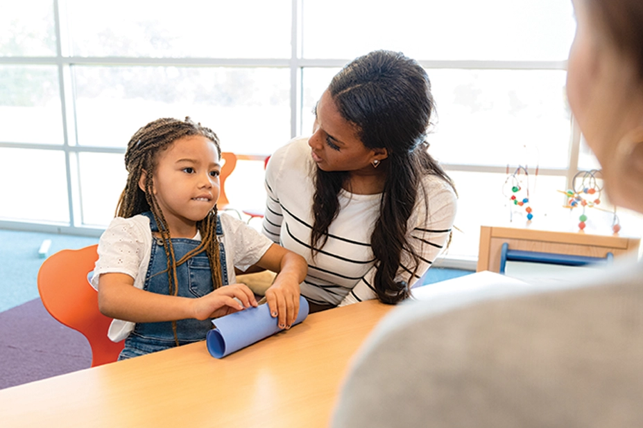 Woman talks with preschool-aged girl at a table in an early childhood classroom.