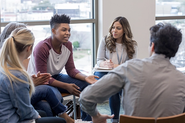 A small group of high school students sit in chairs in a circle in a school library, talking.