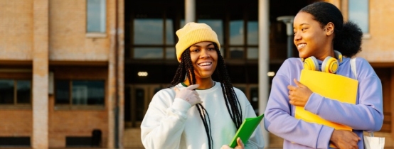 Two teen girls walk together outside a school building, talking and smiling.